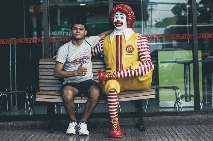A young man sitting on a bench next to a seated Ronald McDonald statue outside a McDonald's restaurant.
