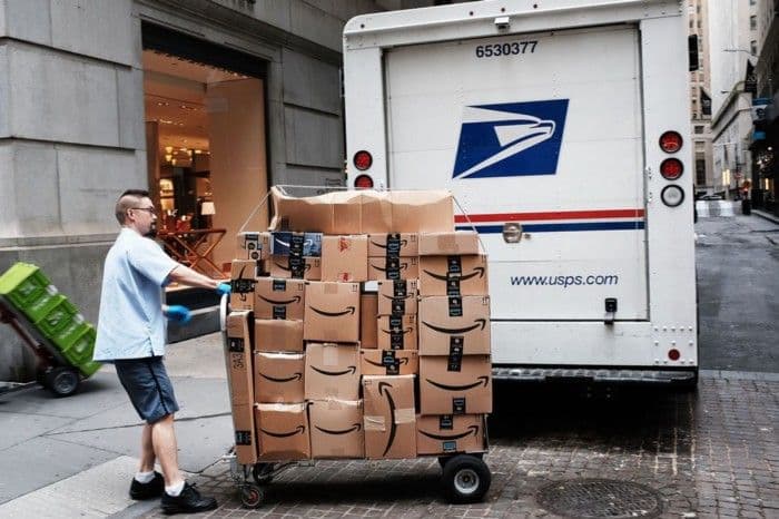 A delivery worker pushes a cart stacked with Amazon boxes past a USPS mail truck on a city street.