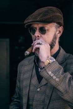 A well-dressed man in a tweed flat cap, sunglasses, and a checked three-piece suit smoking a cigar.