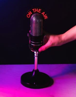 A vintage broadcast microphone on a stand lit by pink and purple light, with a glowing red On The Air sign behind it.