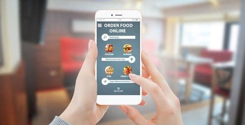 A person holding a smartphone showing a food ordering app with menu categories, in front of a blurred restaurant dining room.