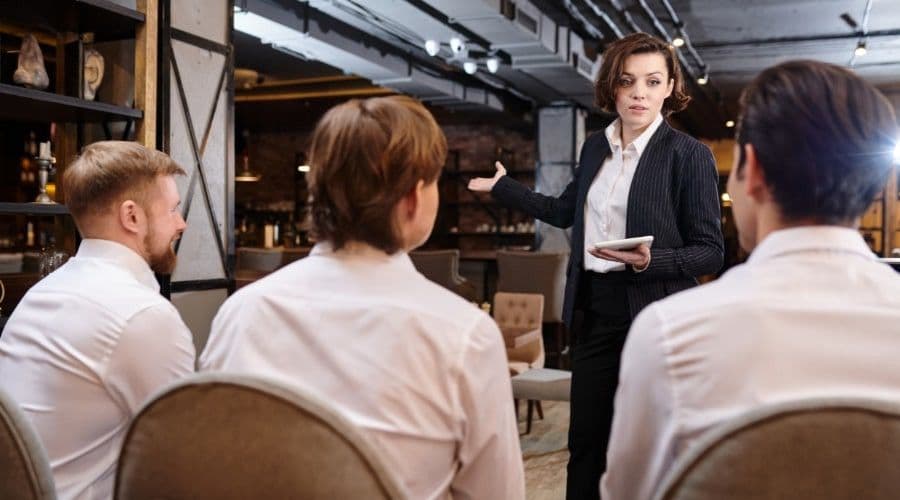 A manager in a blazer leading a training session for three seated servers in a dimly lit restaurant.