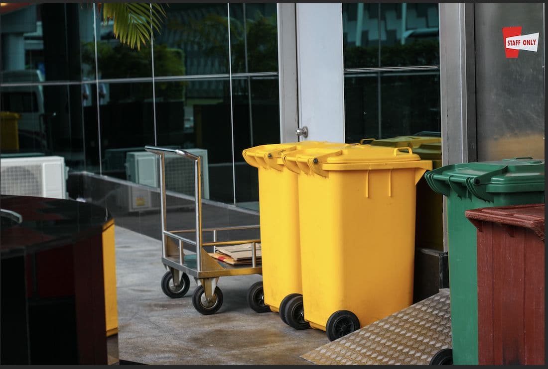 A yellow commercial waste bin and recycling carts parked at a restaurant's back entrance.