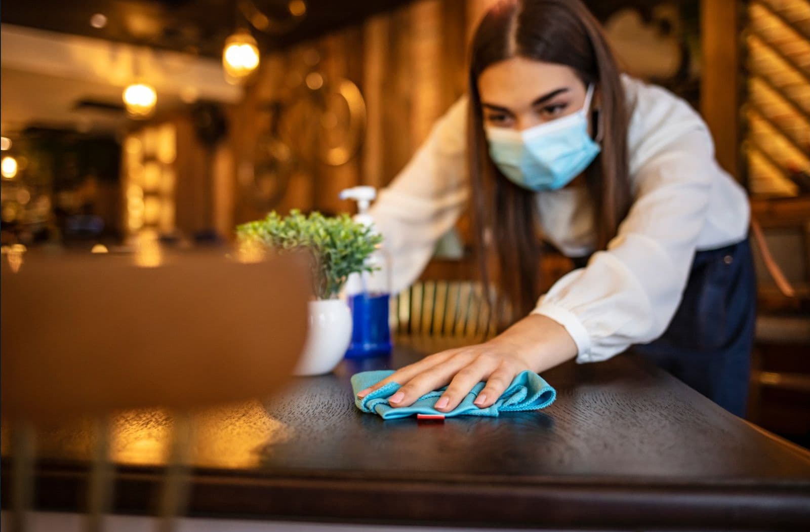 A restaurant worker in a face mask wiping down a wooden bar counter with a cloth and spray bottle.