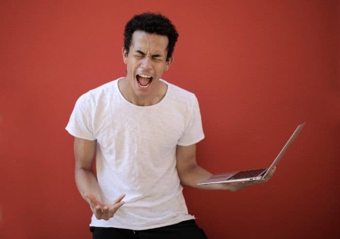 A man in a white shirt holding a laptop and shouting in frustration against a red wall