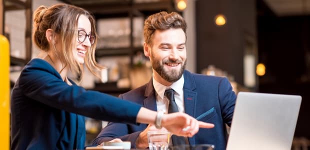 Two colleagues smiling and pointing at a laptop screen in a cafe.