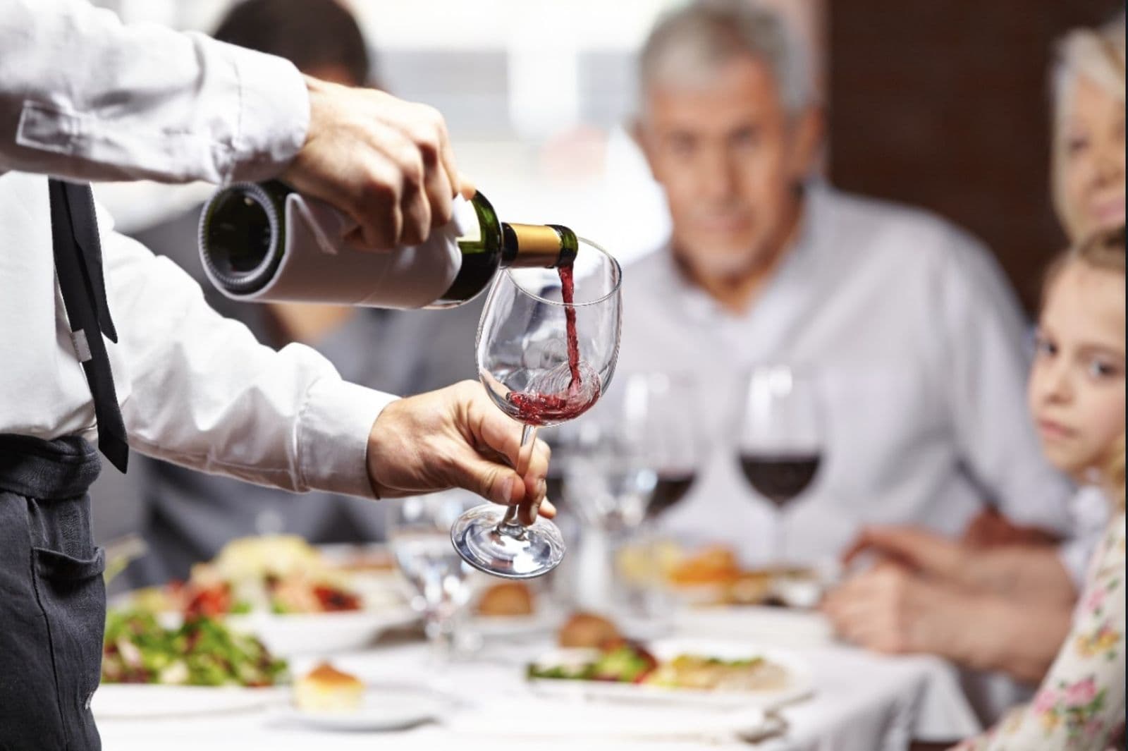 A server in a white shirt pouring red wine into a glass at a restaurant table with seated guests.