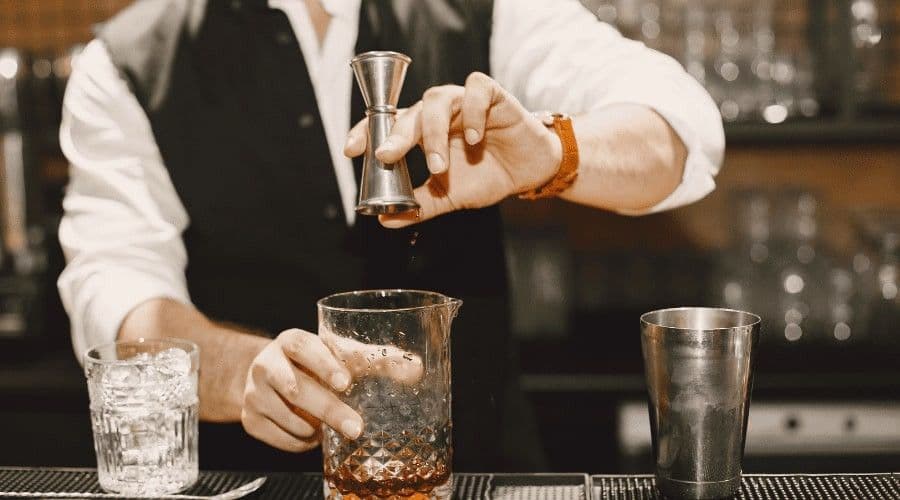 A bartender in a vest measuring a spirit with a jigger into a mixing glass on a bar counter.