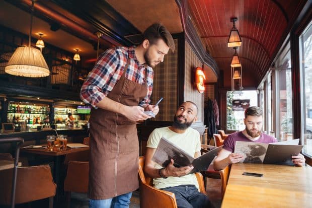 A restaurant server in an apron taking an order on a tablet from two guests reading menus at a table.