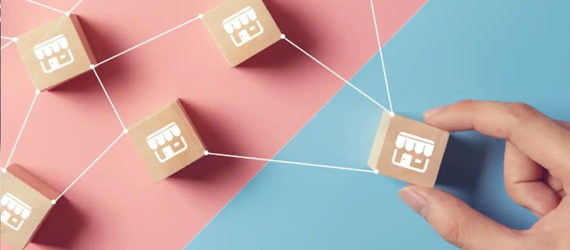 Wooden blocks printed with storefront icons connected by lines, with a hand placing one block, representing a franchise network.