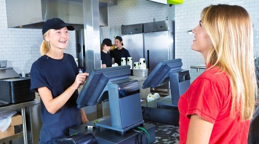 A restaurant employee in a black cap and shirt takes an order at a point-of-sale terminal from a smiling customer in a red shirt.