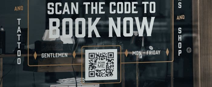 A storefront window decal reading Scan the code to book now, with a printed QR code in the center.