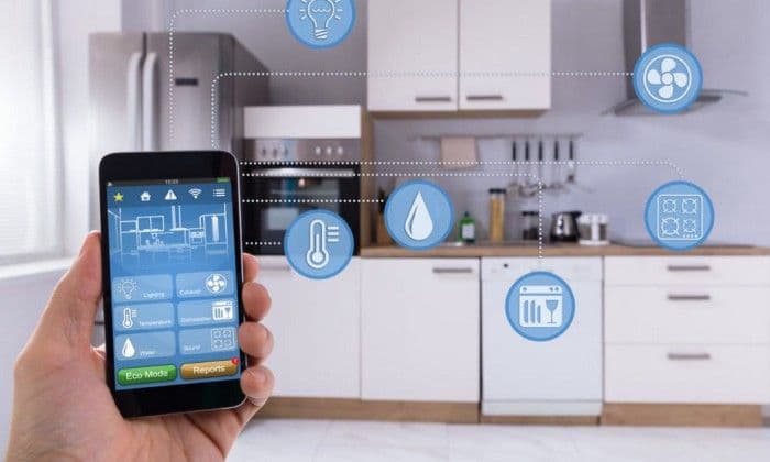 A hand holding a smartphone running a smart home app in front of a modern kitchen, with connected-device icons for lighting, temperature, and appliances.
