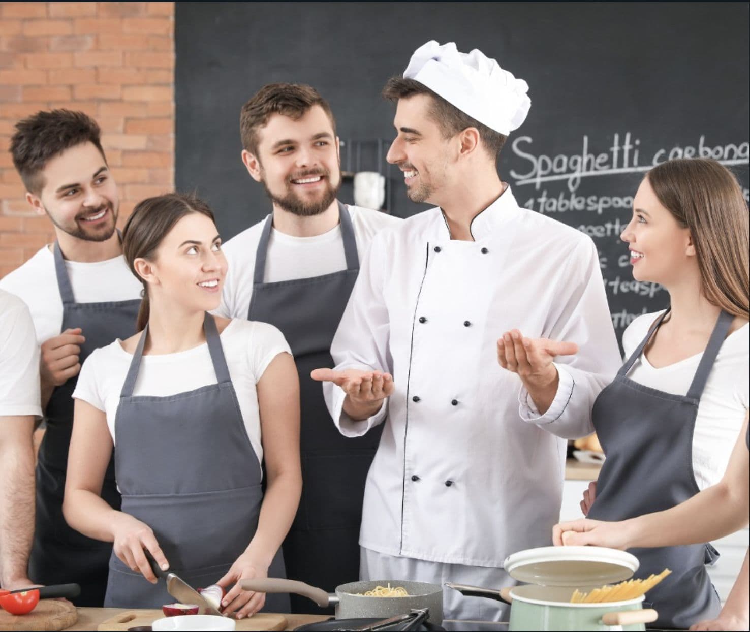 A chef in a white uniform talking and laughing with five restaurant staff in gray aprons during a kitchen training session.