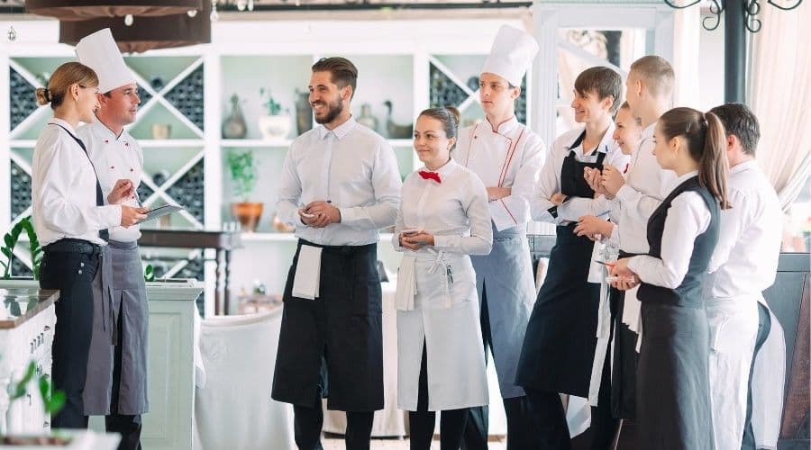A line of restaurant staff in chef whites and aprons standing attentively in a dining room as a manager addresses them.