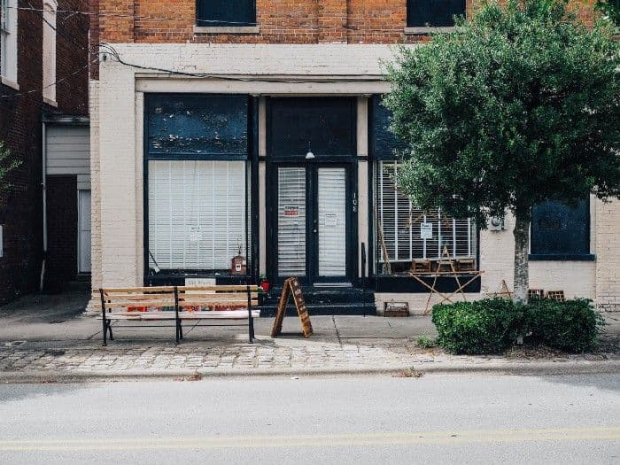 A vacant brick-and-mortar storefront with papered windows, an empty bench, and a sandwich board on the sidewalk.