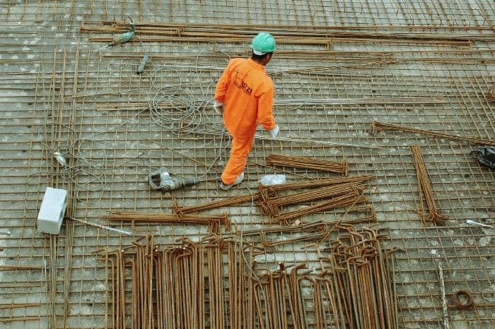 A construction worker in orange coveralls and a hard hat walks across a grid of steel rebar at a building site.