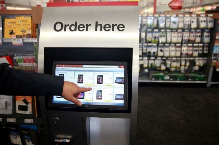 A customer's hand uses a self-service touchscreen ordering kiosk labeled Order here in an electronics store.