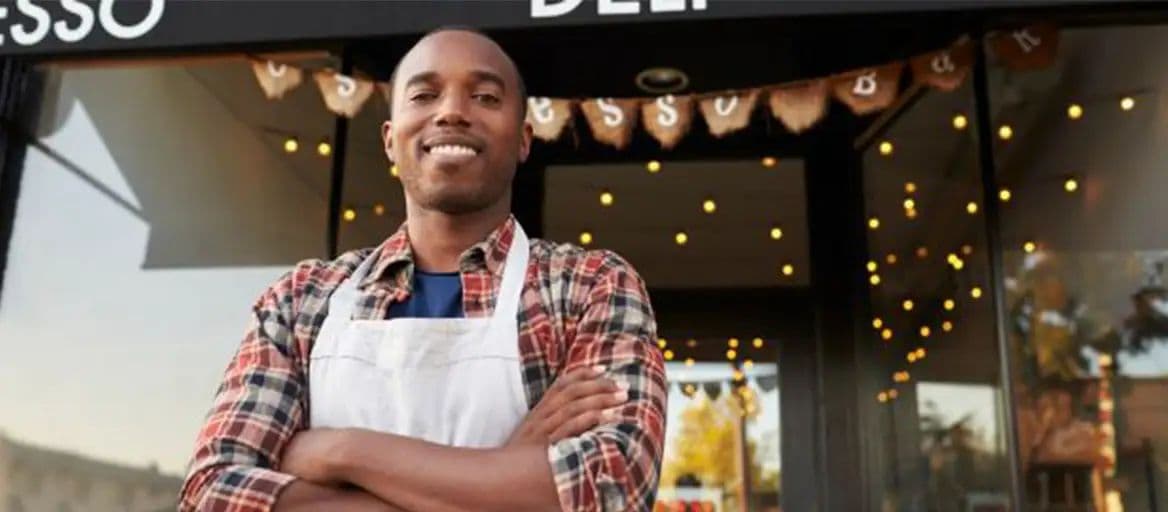 A smiling cafe owner in an apron stands with arms crossed outside his coffee shop storefront.
