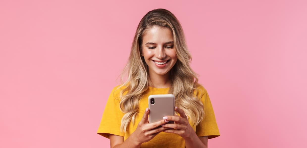 A smiling person in a yellow shirt using a smartphone against a pink background.