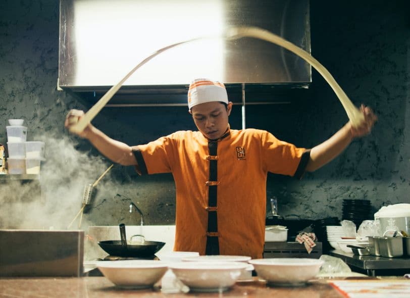 A cook in a white hat stretching dough over steaming bowls in a busy commercial kitchen.