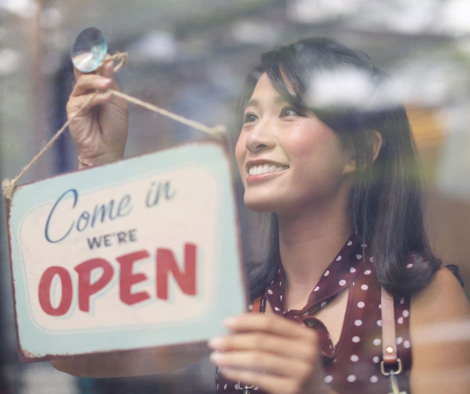 A smiling restaurant owner hanging a “Come in, we’re open” sign in the front window.
