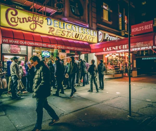 A busy city street at night with diners and pedestrians gathered outside a brightly lit restaurant.