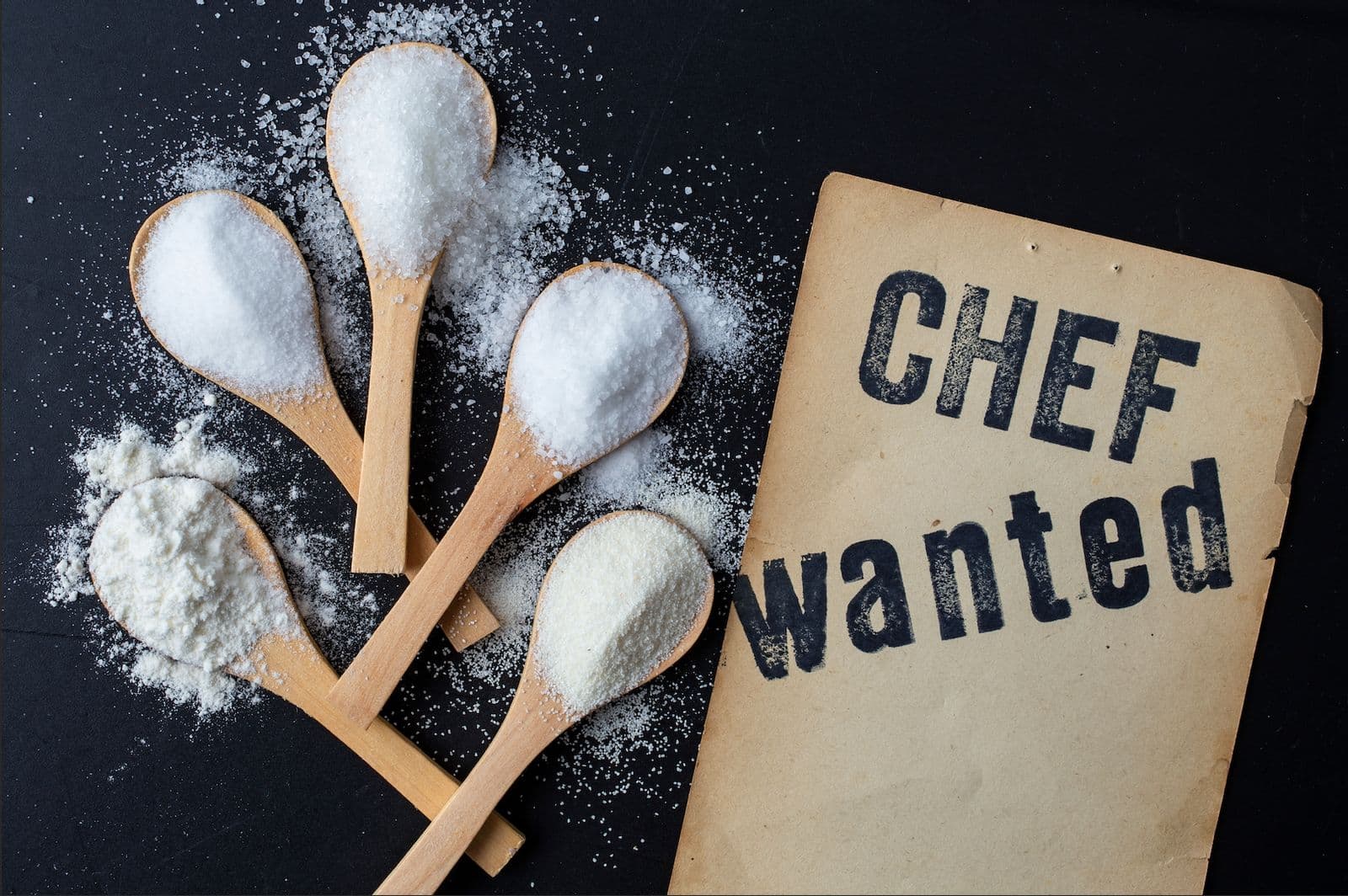Wooden spoons holding flour, sugar, and salt next to a worn paper sign reading CHEF wanted on a dark background.