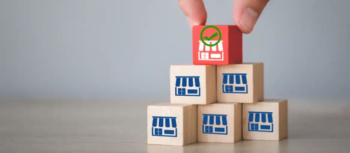 A hand placing a red wooden block with a green checkmark and storefront icon on top of a pyramid of wooden blocks marked with blue storefront icons.