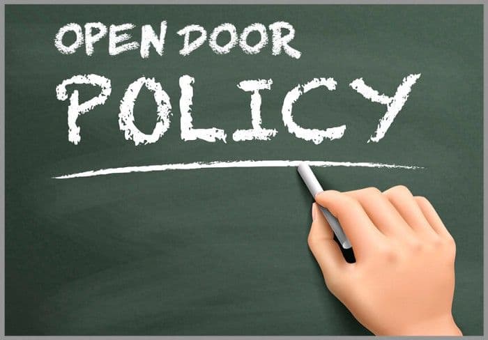The words Open Door Policy written in white chalk on a green chalkboard, with a hand holding a piece of chalk
