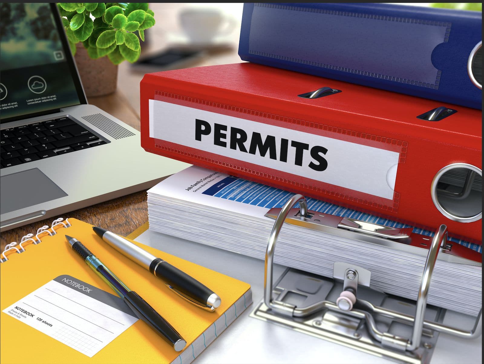 A red binder labeled PERMITS sitting on a stack of paperwork next to a laptop, notebook, and pens on a desk.