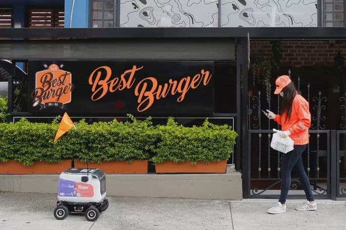 A masked delivery worker walks past a Best Burger storefront as a sidewalk delivery robot rolls by.