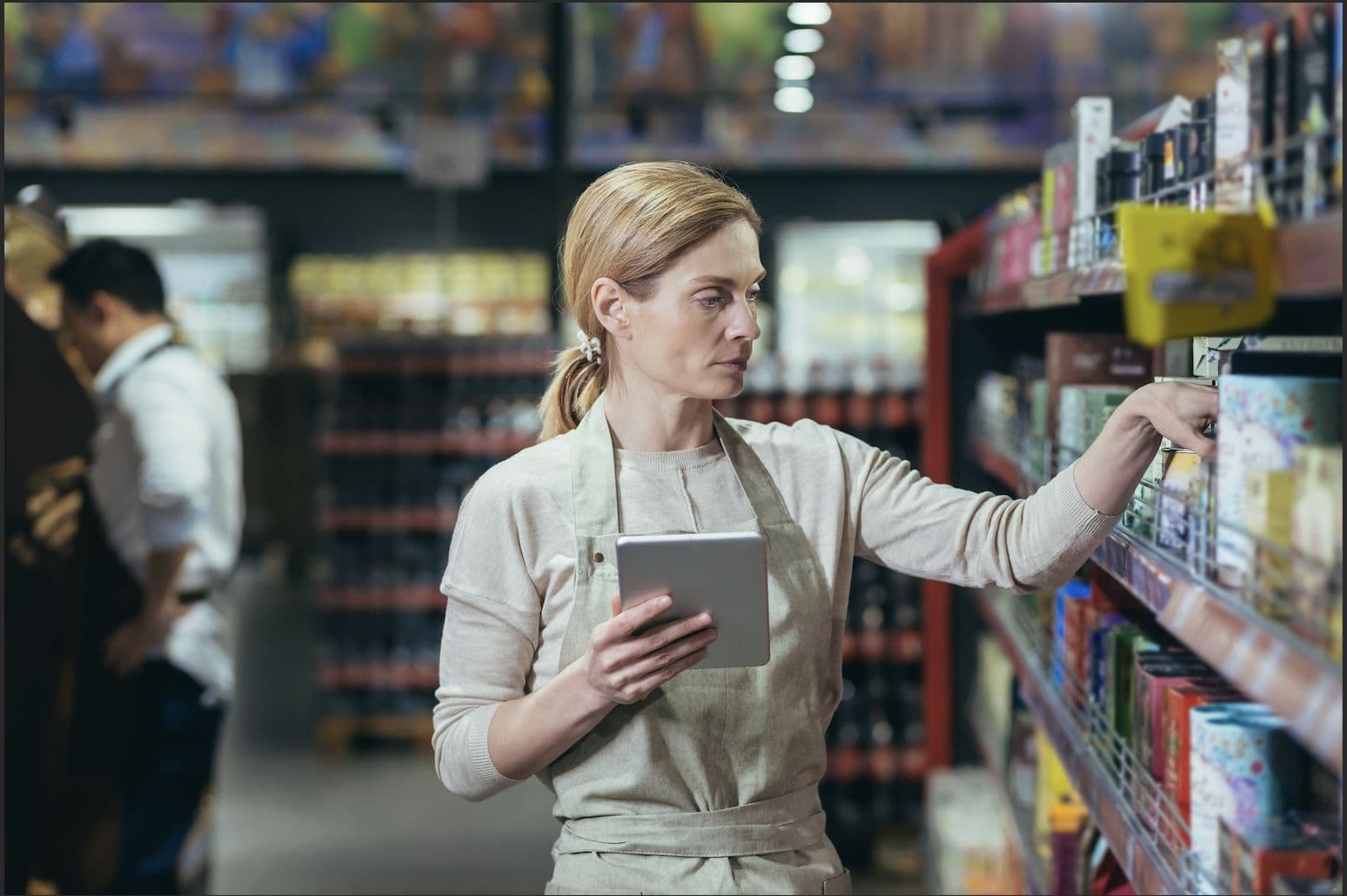 A restaurant worker in an apron checks shelf stock with a tablet in a stockroom aisle.