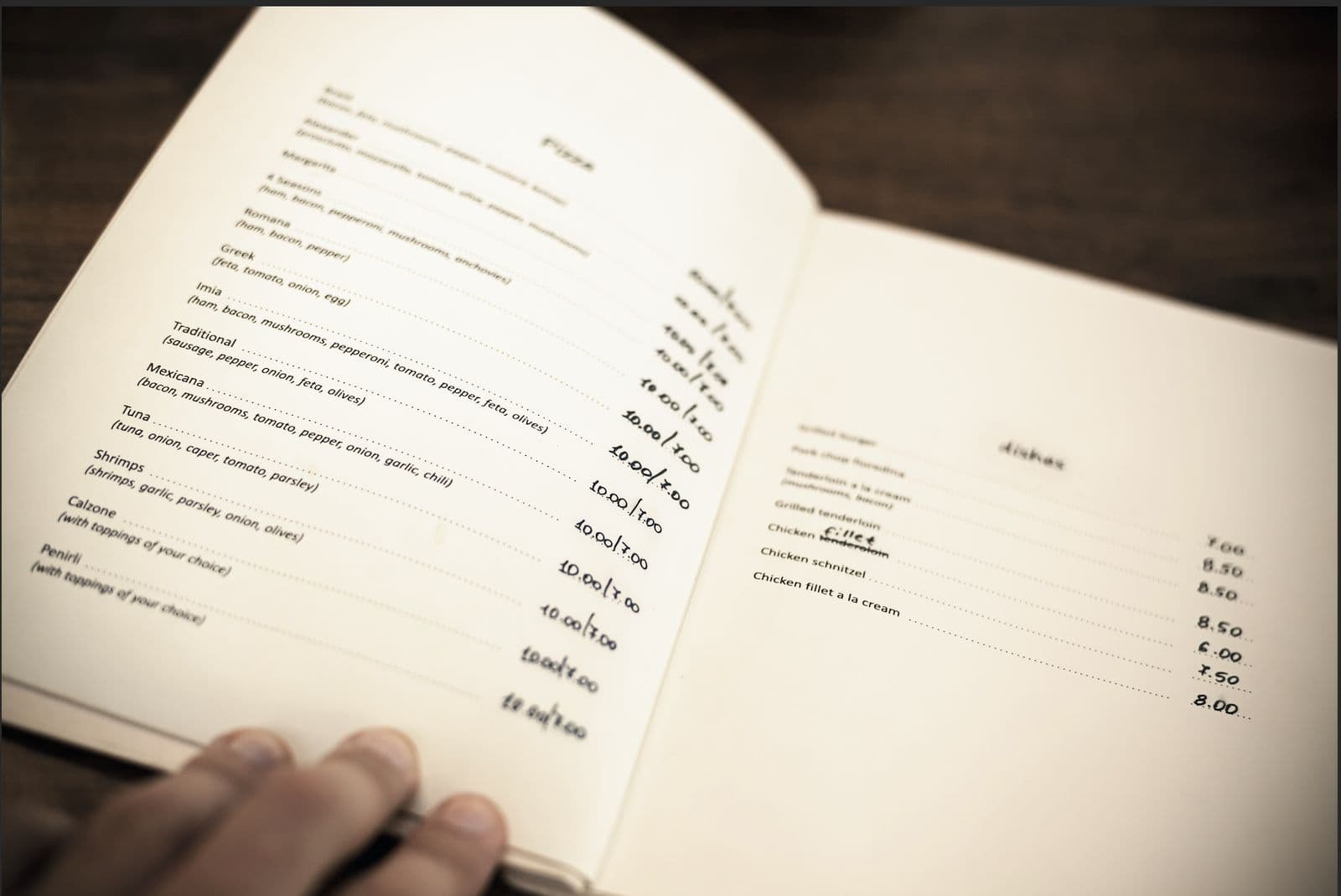 An open printed restaurant menu showing handwritten dish names and prices, held open by a hand on a dark wooden table.