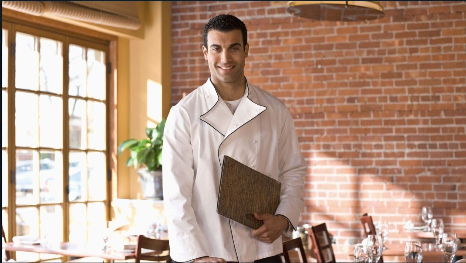 A smiling restaurant manager in a white chef jacket holding a menu board in a brick-walled dining room.