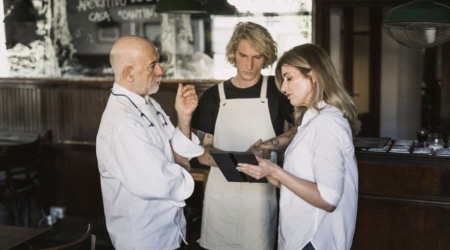 A senior chef, a cook in an apron, and a manager reviewing notes on a tablet together in a restaurant.