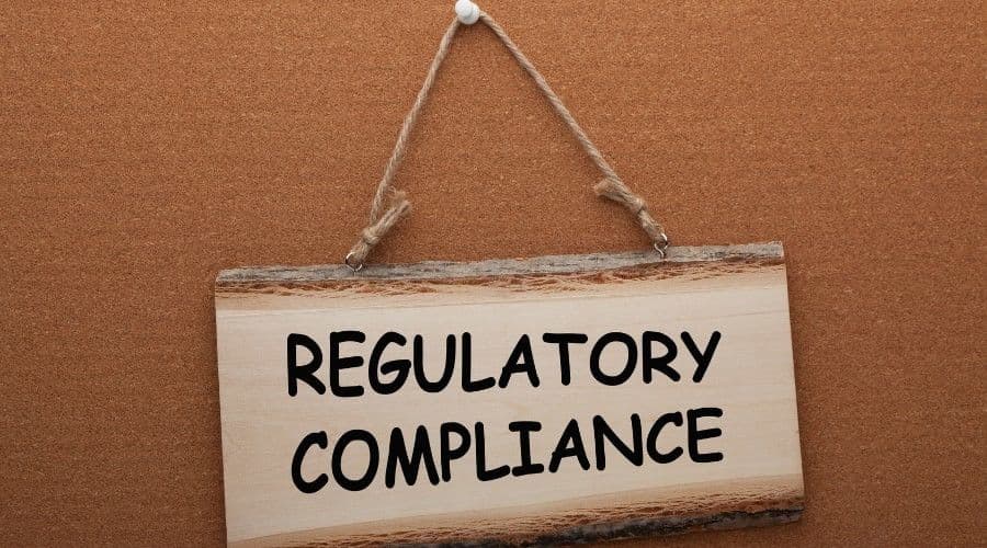 A wooden sign reading Regulatory Compliance hanging by twine against a cork board.