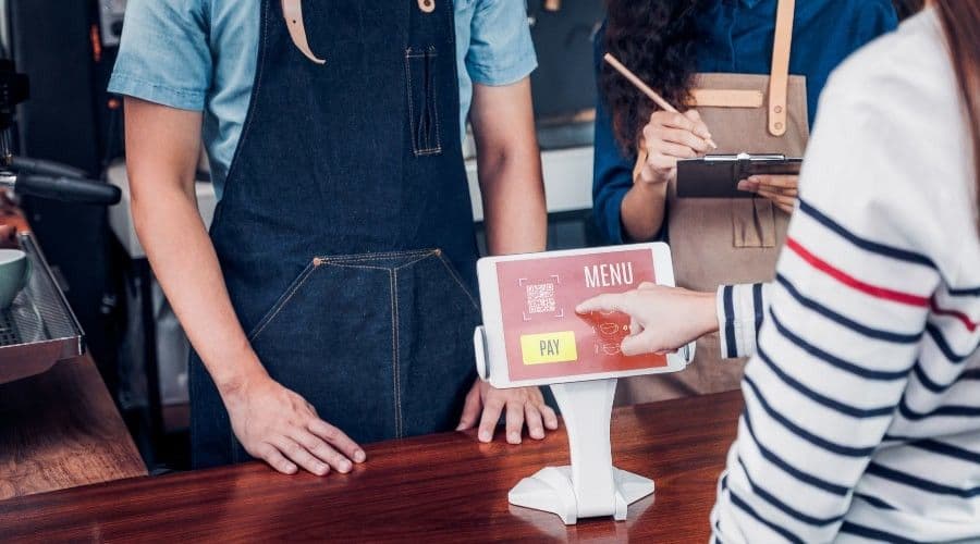 A customer taps the touchscreen of a countertop self-ordering kiosk at a cafe while two staff members in aprons stand behind the counter.