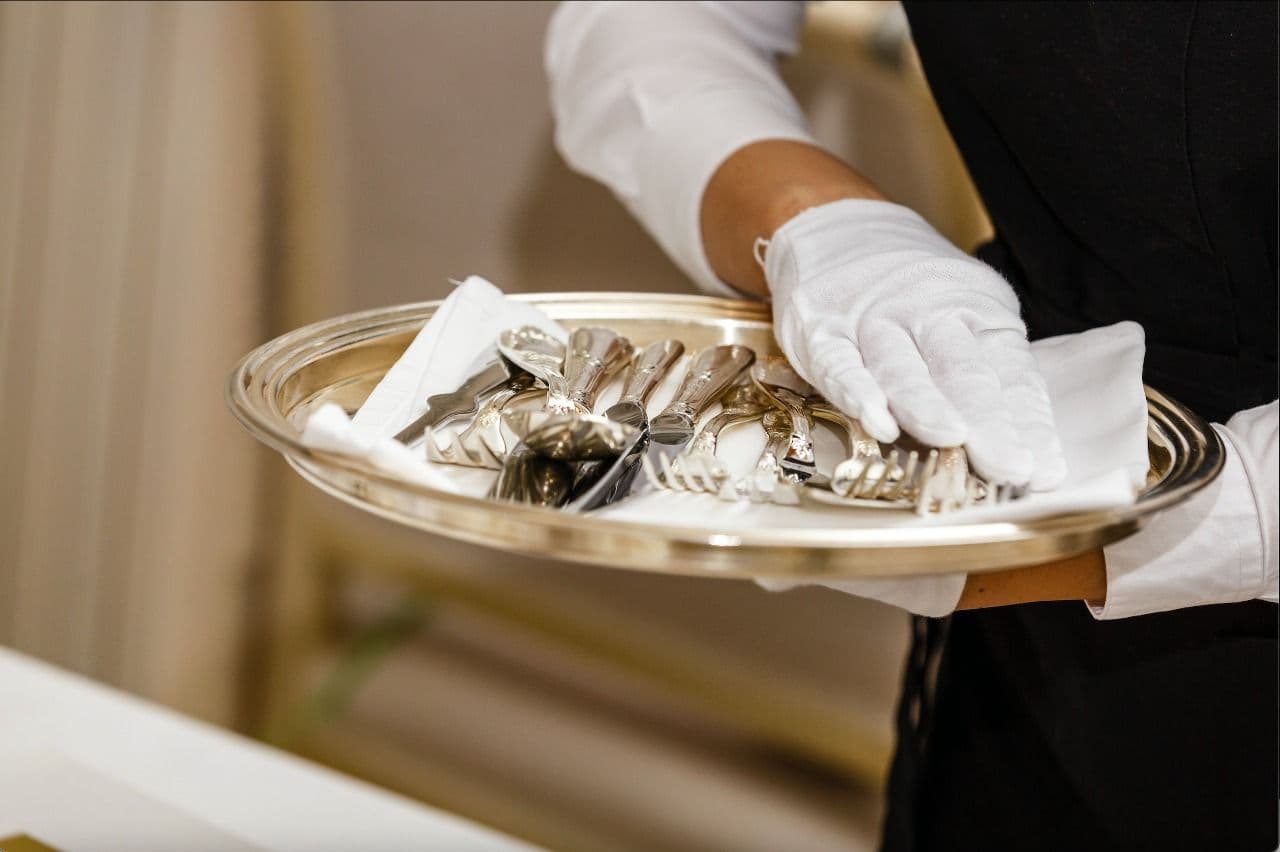 A server in a black uniform and white gloves carrying a gold tray of polished silverware and a folded napkin.