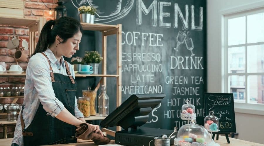 A barista in an apron wipes down a counter beside a point-of-sale terminal under a chalkboard coffee menu.
