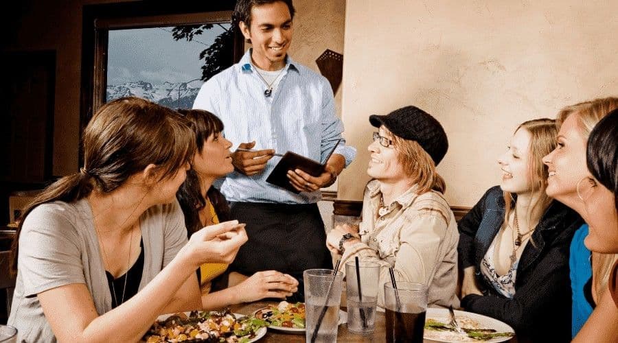 A smiling server taking an order from a group of guests seated around a restaurant table.
