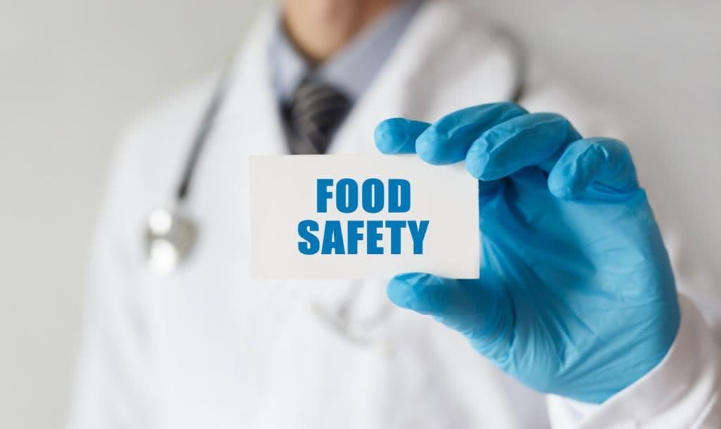A person in a white coat and blue glove holding a card that reads Food Safety.