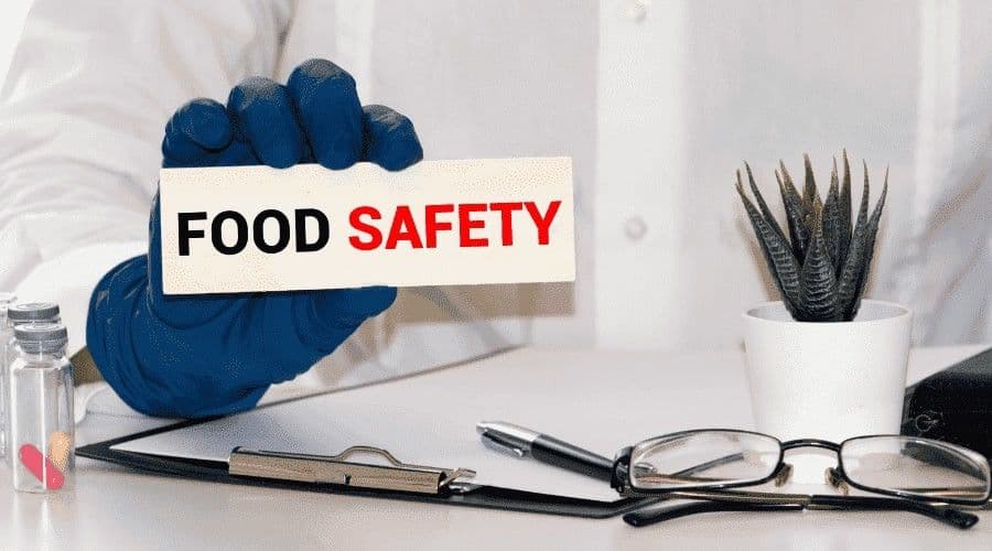 A gloved hand holding a Food Safety card over a desk with a clipboard, pen, glasses, and a small plant.