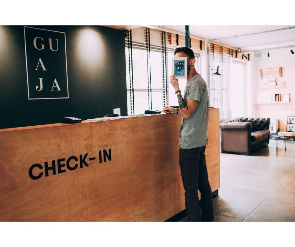 An hourly worker in a gray t-shirt standing at a wooden CHECK-IN desk in a modern lobby, holding a card up to his face.