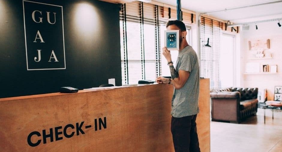 Wide view of an hourly worker standing at a wooden CHECK-IN reception desk in a bright lobby with leather seating in the background.