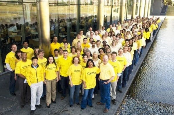 A large group of company employees in matching yellow shirts gathered outside a glass office building.
