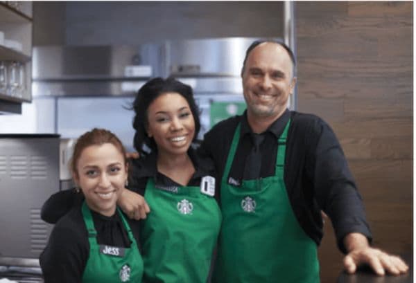 Three smiling Starbucks baristas in green aprons standing together behind the counter.