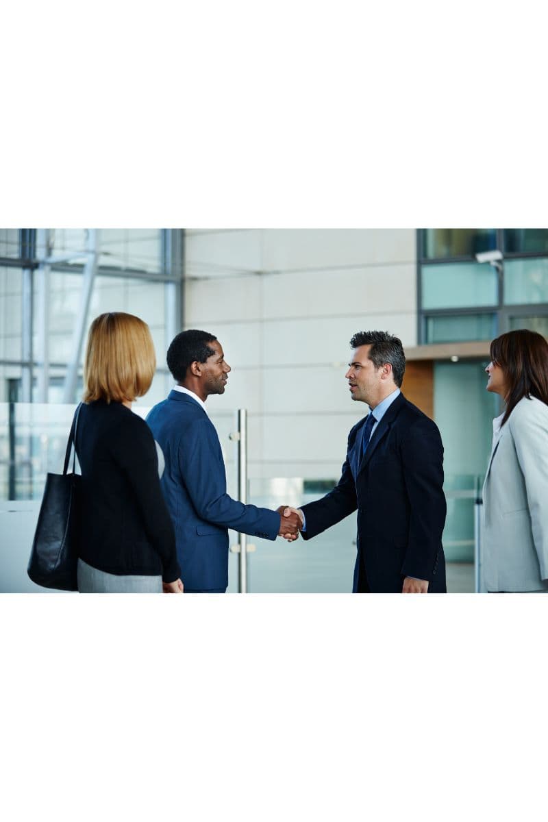 Two business professionals shaking hands in an office lobby while two colleagues look on.