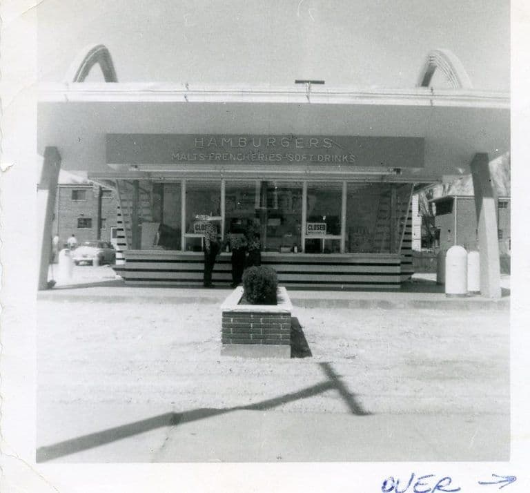 A vintage black-and-white photo of an early McDonald's hamburger stand with the original double arches and a Hamburgers sign.