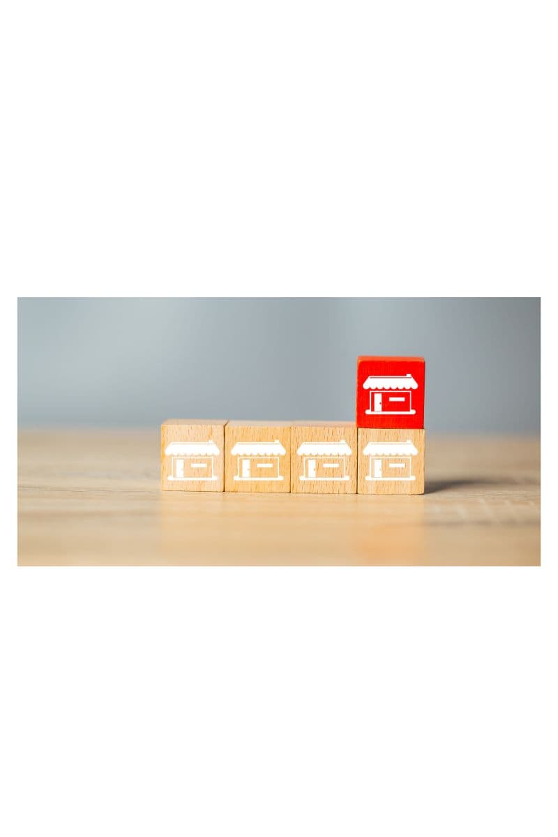 Four wooden blocks printed with white storefront icons on a desk, with one block flipped to a red storefront face standing apart from the row.